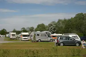 Campingplatz mit Wohnmobilen und Autos auf Stellplätzen, Rasenfläche und Bäumen im Hintergrund.