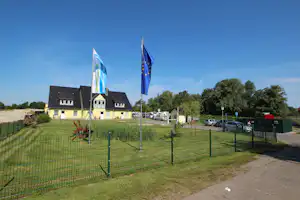 Campingplatz-Gebäude mit zwei Fahnen vor dem Eingang, Rasenfläche, Autos und Wohnwagenstellplätze im Hintergrund.