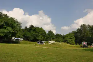 Wohnmobil-Stellplatz auf einer Wiese mit Wohnwagen und Bäumen, im Hintergrund ein Wohnmobil unter blauem Himmel mit Wolken.