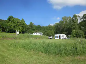 Mehrere Wohnwagen und ein Wohnmobil stehen auf einer grünen Wiese am Campingplatz, umgeben von Bäumen.