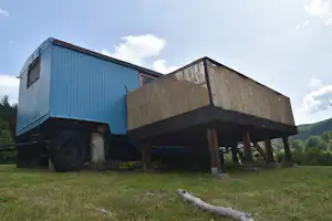 Holzunterkunft auf Stelzen neben einem blauen Container, auf einer Wiese mit Zaun und Holzstamm im Vordergrund.