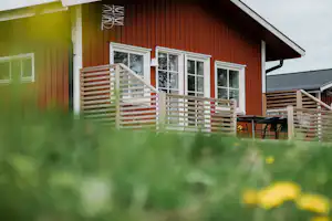 Holzverkleidetes Ferienhaus mit großen Fenstern und Holzterrasse, davor ein Zaun und Gartenmöbel auf dem Campingplatz.
