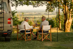 Camping-Van am Stellplatz, zwei Klappstühle am Tisch unter einer Markise, Laternen und Blick ins Tal durch Bäume.