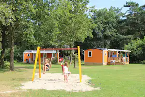 Oranger Wohnwagen und Spielplatz mit gelbem Klettergerüst im Grünen, daneben Gartenmöbel und Bäume.