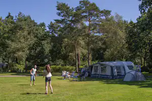 Campingwiese mit Wohnwagen und Vorzelt, daneben Zelte und Campingstühle unter Bäumen im Grünen.