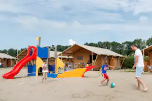 Spielplatz auf dem Campingplatz mit großer Rutsche, Sandfläche und Wohnzelten im Hintergrund, Kinder spielen.