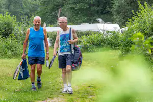 Zwei Camper laufen auf einem Grasweg im Grünen, mit Rucksäcken und Trinkflasche, im Hintergrund ein Wohnwagen und Zelt.