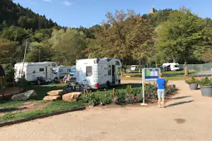 Wohnmobile auf dem Campingplatz, gepflasterter Weg mit Blumenbeeten und Informationstafel, im Hintergrund Wald und Bäume.