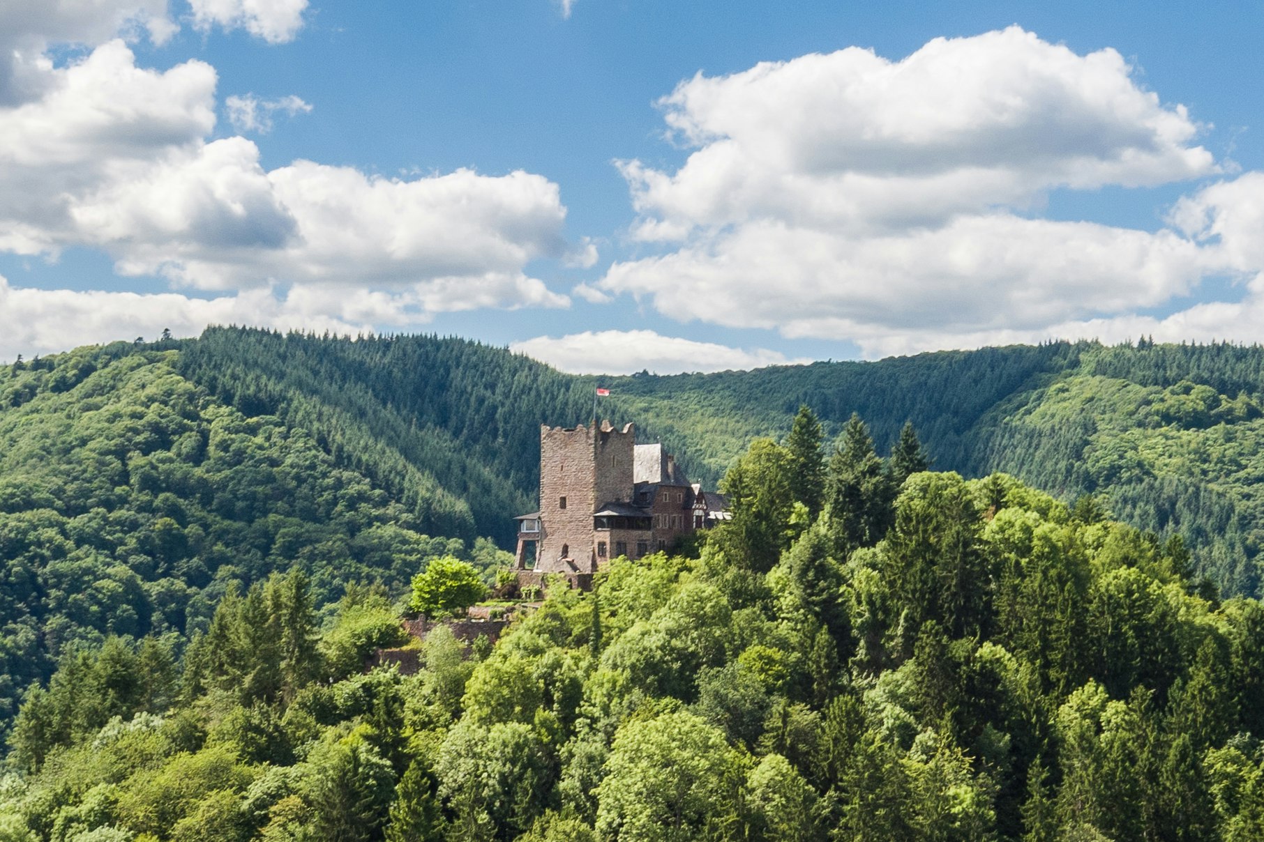 Burganlage im grünen Waldtal, umgeben von Bäumen und Hügeln, mit blauem Himmel und Wolken.