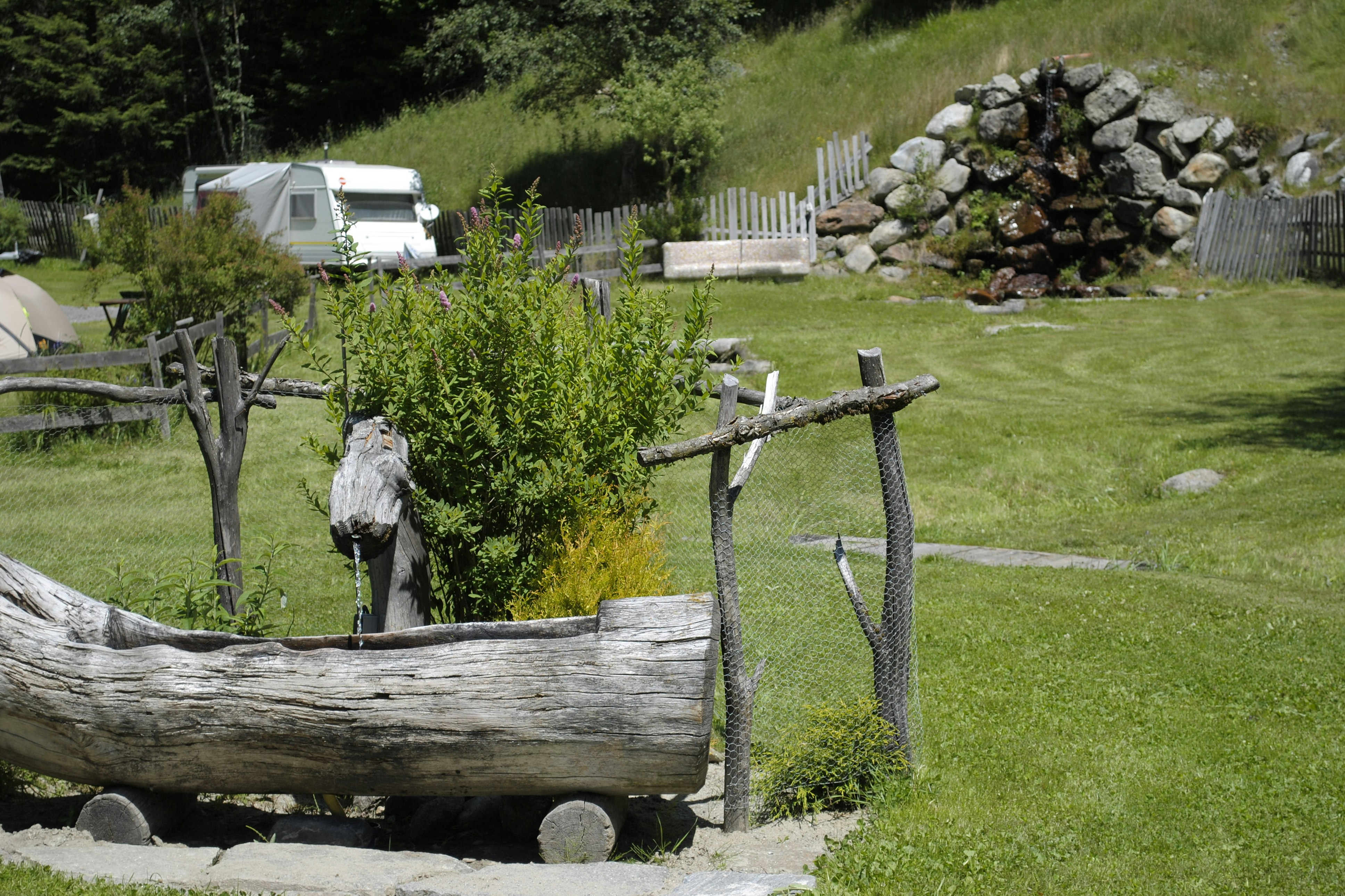 Grüner Campingplatz mit Rasen, Holzgeländer und einem kleinen Gartenweg; im Hintergrund ein Wohnwagen und ein Steinhaufen.