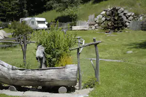 Grüner Campingplatz mit Rasen, Holzgeländer und einem kleinen Gartenweg; im Hintergrund ein Wohnwagen und ein Steinhaufen.