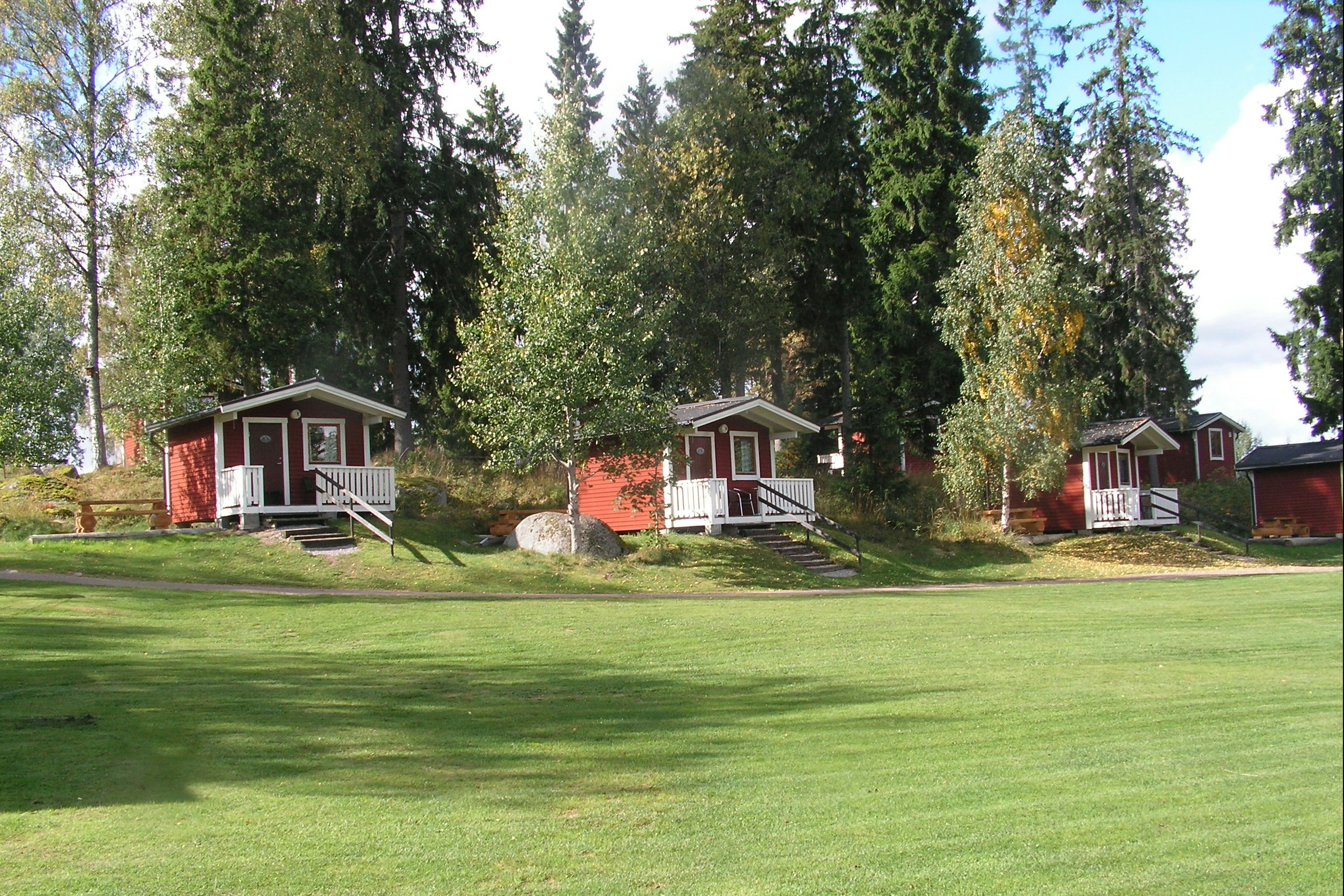 Mehrere rote Ferienhütten mit Veranda auf einem gepflegten Rasen, umgeben von hohen Bäumen im Campingbereich.