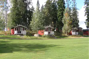 Mehrere rote Ferienhütten mit Veranda auf einem gepflegten Rasen, umgeben von hohen Bäumen im Campingbereich.