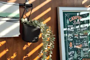 Blumentopf mit Sukkulenten an einer Camping-Wand, daneben ein gerahmtes Schild und ein Metallkorb auf dem Tisch.