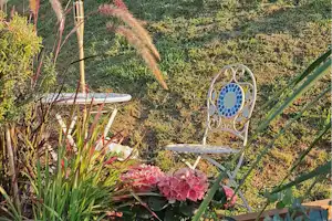 Blumenbeet mit Holzkiste und pinken Blüten, dahinter eine Terrasse mit Holzgeländer und Sonnenschirmstangen vor blauem Himmel.