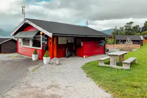 Rote Holzhütte mit überdachter Terrasse, Sitzbank und Tisch aus Holz, Kiesweg und grüner Rasenfläche am Campingplatz.