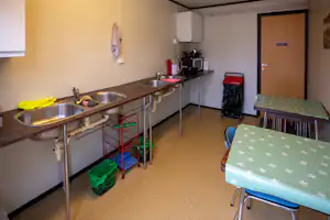 Gemeinschaftsküche im Campingbereich mit Edelstahlspuelen, Arbeitsplatte, Tisch und Stühlen sowie Mülleimer und Tür im Hintergrund.