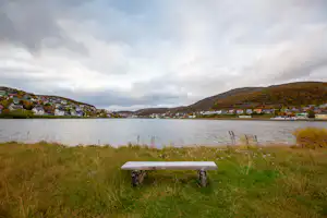 Picknicktisch auf einer Wiese am See, im Hintergrund Ufer mit Häusern und Hügeln unter bewölktem Himmel.