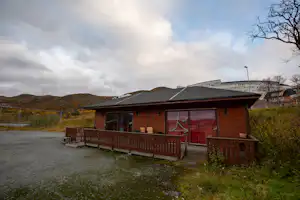 Holzbungalow mit Terrasse und roten Fenstern auf einem Campingplatz, im Hintergrund Gebäude und Hügel.