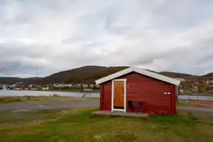 Rote Holzhütte auf einer Wiese am Campingplatz, mit See im Hintergrund und Hügeln unter bewölktem Himmel.