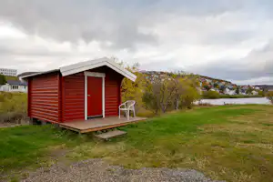 Holzhaus auf einem Campingplatz mit roter Fassade, kleiner Veranda und Treppe, umgeben von Rasen und Hügeln.