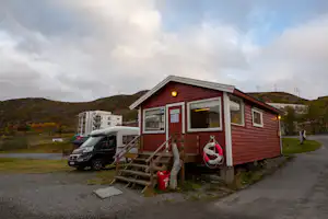Rotes Mobilheim mit Holztreppe, davor ein Wohnmobil und ein geparkter Transporter am Weg, im Hintergrund Berge und Wiese.