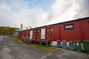 Rotes Campinghaus mit Terrasse, Stufen und Geländer, daneben Mülltonnen und ein Kiesweg entlang der Gebäudezeile.