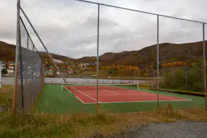 Tennisplatz auf dem Campingplatz, umgeben von einem hohen Zaun, mit Blick auf Hügel und Häuser im Hintergrund.