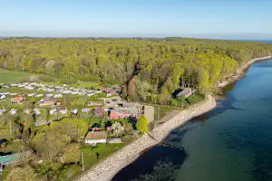 Campingplatz am Seeufer mit Wohnwagen und Zelten, schmaler Kiesstrand, umgeben von Wald und einem Weg entlang des Wassers.
