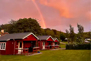 Rote Campinghäuser mit Veranda und Sitzbereich, umgeben von Rasen und Büschen, im Abendhimmel mit Wolken.