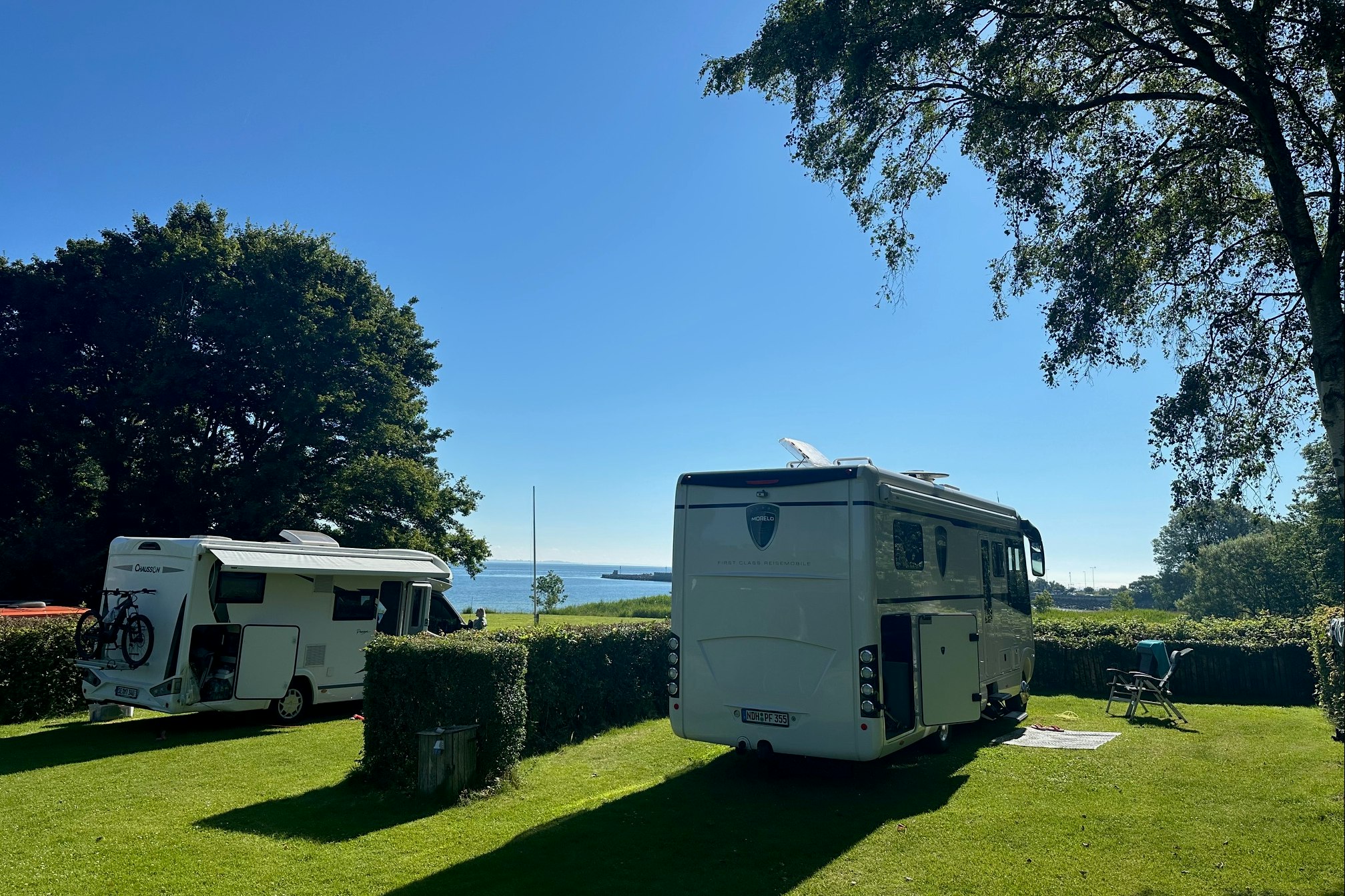 Zwei Wohnmobile auf einem grünen Campingplatz, Hecken und Rasenflächen, im Hintergrund Wiese und Himmel.
