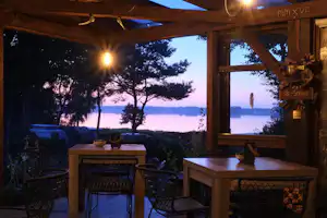Camping-Terrasse am Abend mit Tisch und Stühlen, Lichterkette und Blick auf den See zwischen Bäumen.