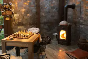 Innenraum einer Holzunterkunft mit Kamin, Schachbrett am Tisch, Stühlen und Sitzbank unter sichtbaren Holzbalken.