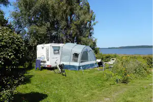 Campingzelt und Wohnwagen auf einem Grasstellplatz, mit Stühlen und Blick auf den See hinter Bäumen.