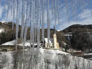 Birken im Vordergrund, dahinter eine Kirche und verschneite Häuser in einem hügeligen Tal.