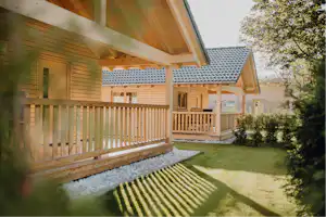 Holzbungalow mit überdachter Terrasse, Holzgeländer und Rasenfläche im Campingbereich, daneben Hecke und Gartenweg.