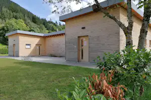 Campinggebäude mit Holzfassade, Eingangstür und Rasenfläche, im Hintergrund bewaldete Berge und ein Nebengebäude.