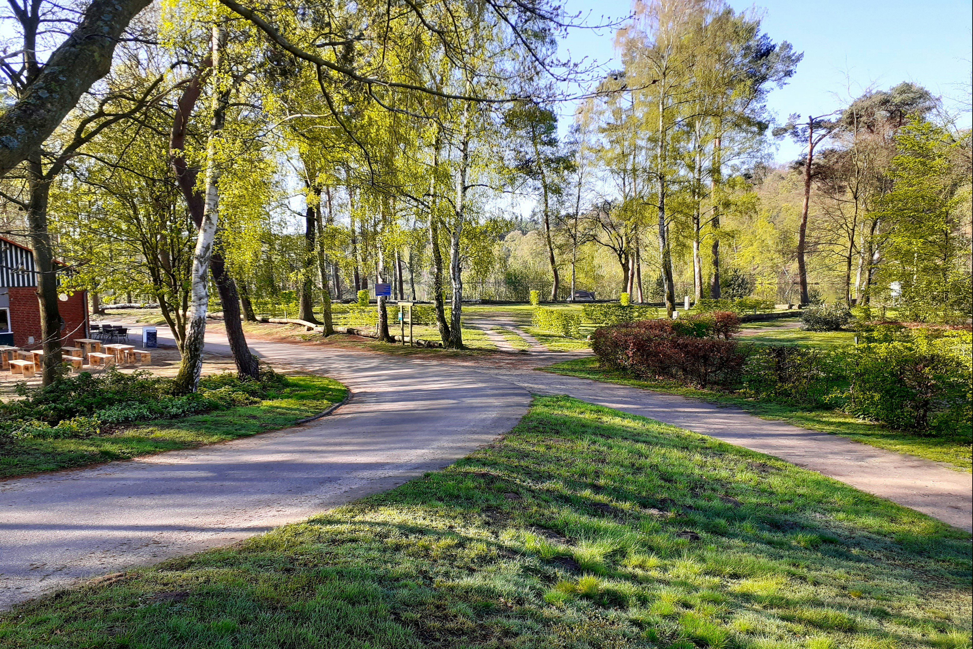 Schotterweg durch einen gepflegten Campingplatz mit Rasenflächen, Büschen und hohen Bäumen, im Hintergrund ein Gebäude.