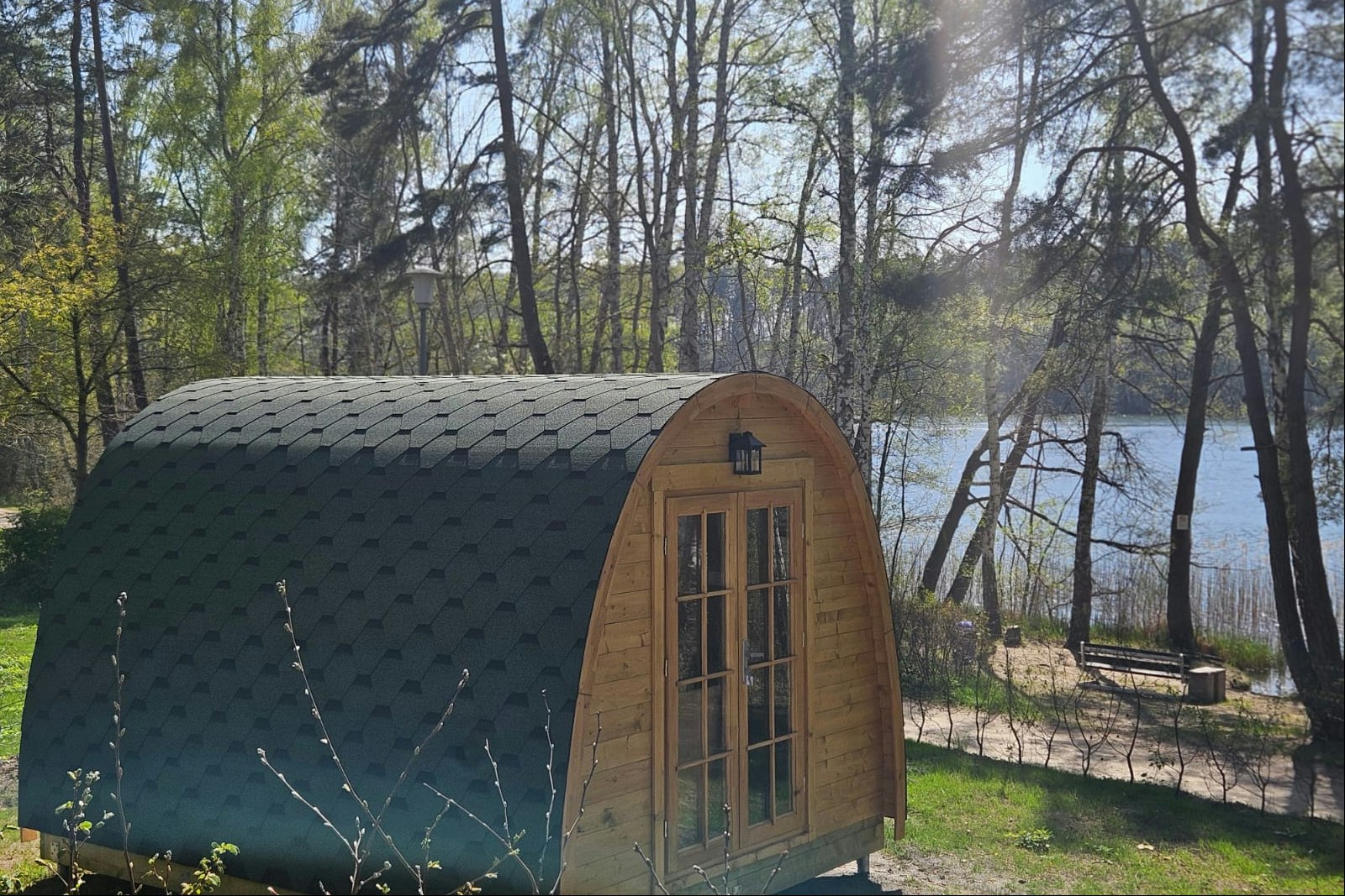 Holz-Zelt mit Fenster und Tür im Campingbereich, umgeben von Bäumen und Gras, im Hintergrund ein Weg und Sitzbank.