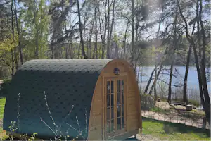 Holz-Zelt mit Fenster und Tür im Campingbereich, umgeben von Bäumen und Gras, im Hintergrund ein Weg und Sitzbank.