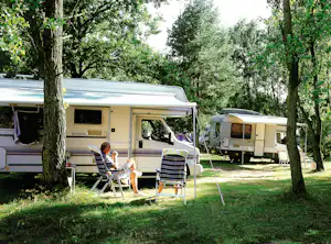 Wohnwagen auf dem Campingplatz unter Bäumen, mit Markise, Stühlen und Tisch vor dem Eingang.