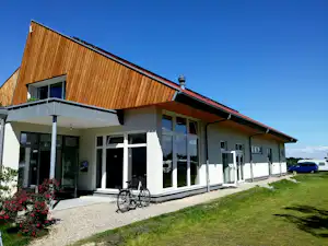 Modernes Holzhaus mit großen Fenstern, überdachter Terrasse und Fahrrad vor dem Eingang, Kiesweg und Rasen im Vordergrund.