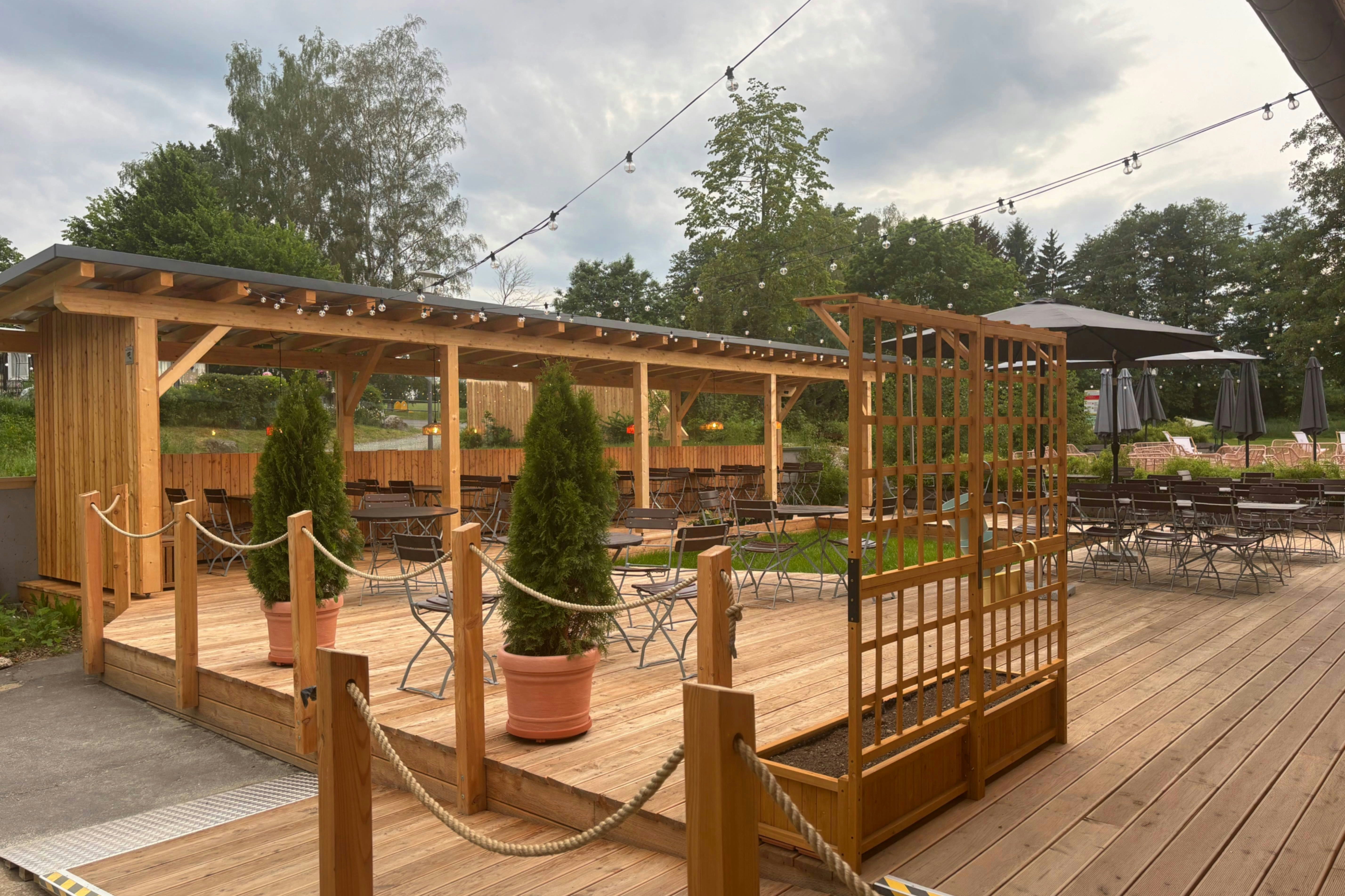 Holzterrasse eines Campingplatzes mit überdachtem Sitzbereich, Seilgeländer, Tischen und Stühlen sowie mehreren Zelten im Hintergrund.