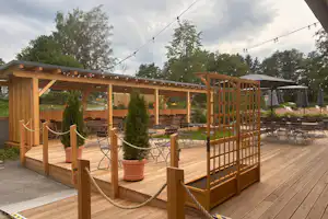 Holzterrasse eines Campingplatzes mit überdachtem Sitzbereich, Seilgeländer, Tischen und Stühlen sowie mehreren Zelten im Hintergrund.
