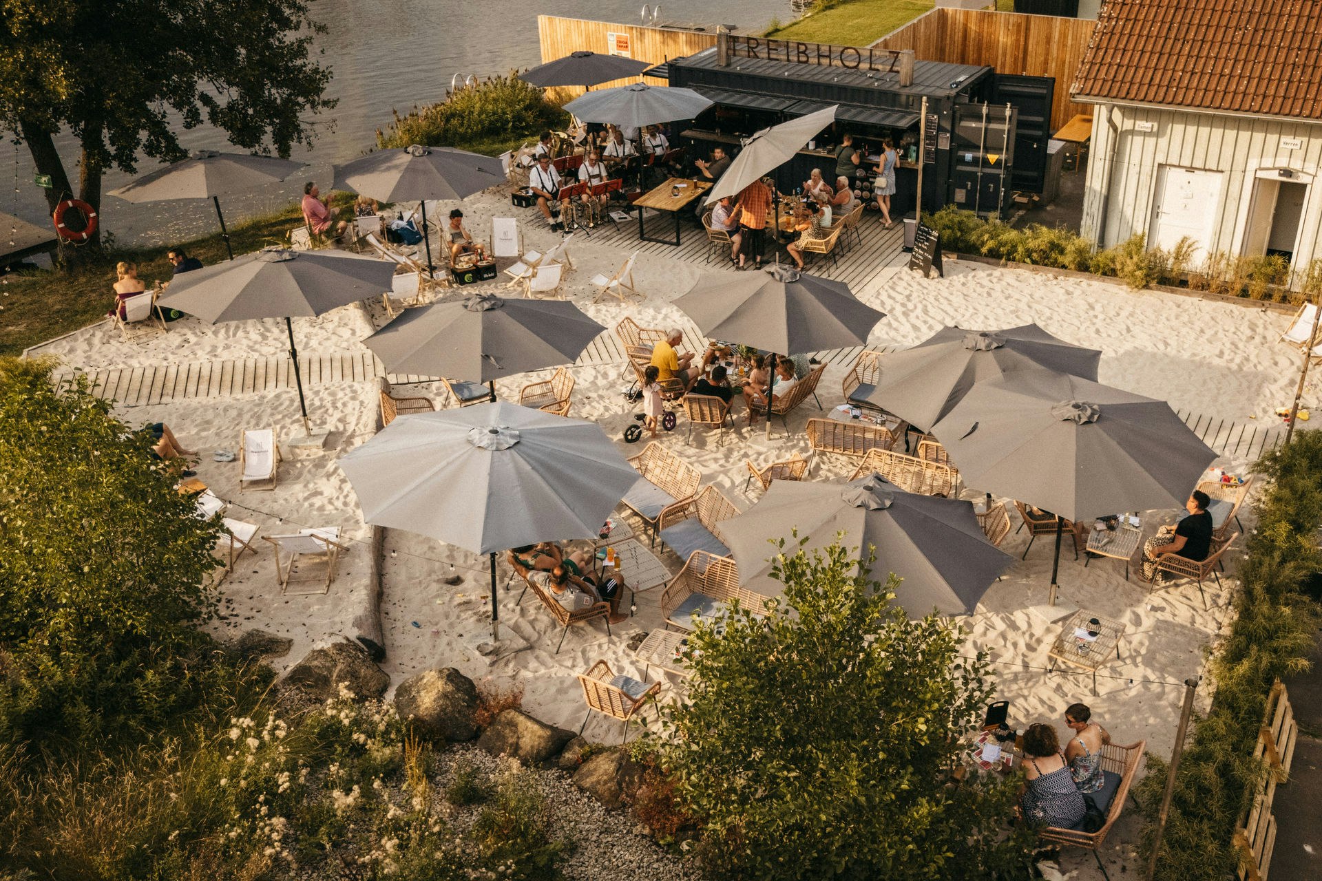 Terrasse auf dem Campingplatz mit mehreren Zelten, vielen Sonnenschirmen und Sitzgruppen auf Kiesboden.