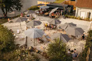 Terrasse auf dem Campingplatz mit mehreren Zelten, vielen Sonnenschirmen und Sitzgruppen auf Kiesboden.