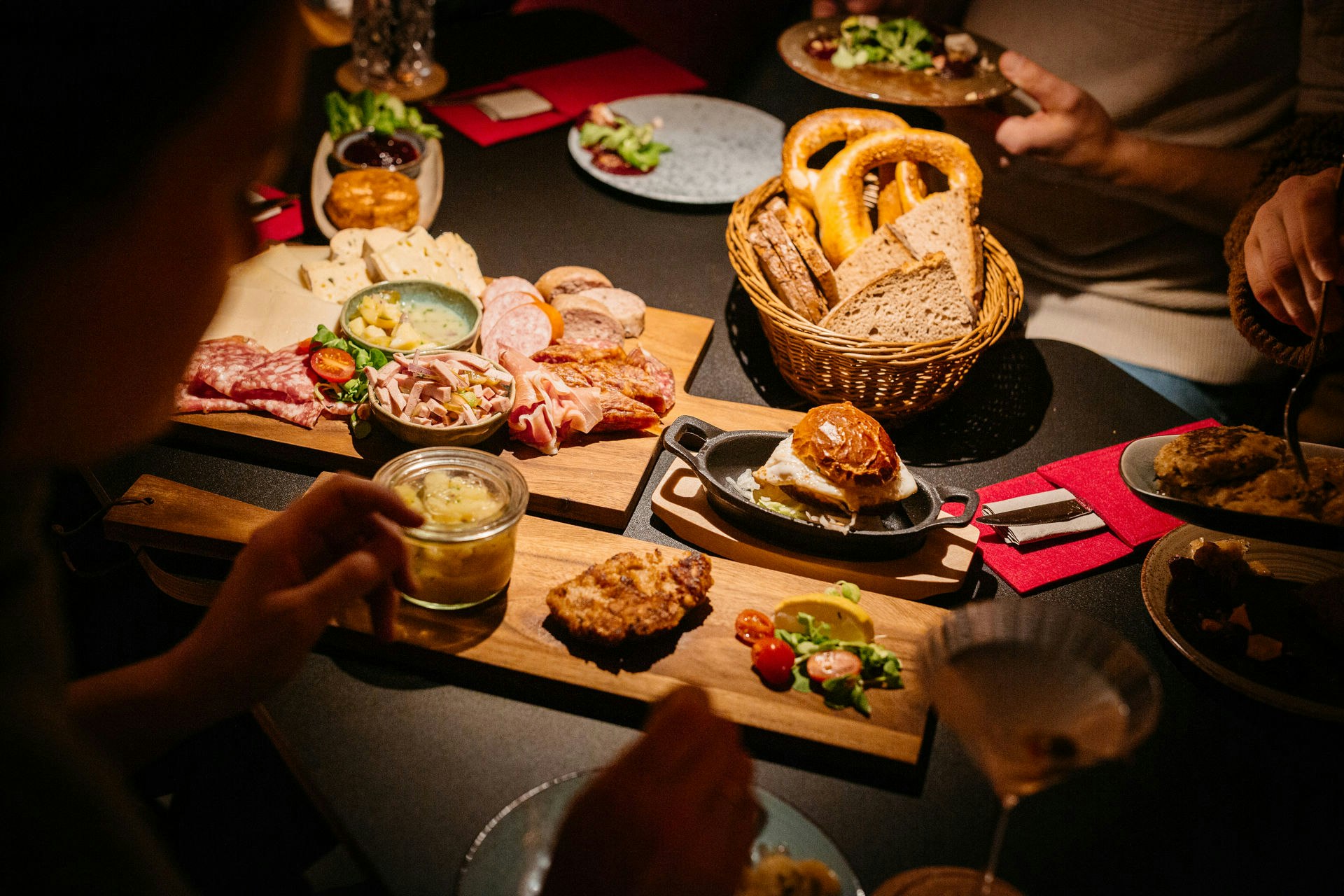 Camping-Tisch mit gegrilltem Essen, Brot im Korb und Tellern, dazu Gläser und Besteck auf einer Holzplatte.