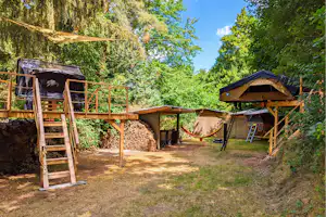 Holzplattform mit Zelt auf einem Campingplatz, daneben Hängematte, Stühle und Wege im Grünen zwischen Bäumen.