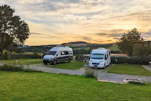Zwei Wohnmobile auf einem Campingplatz, Grasfläche und Kiesweg, Bäume und Hecken im Hintergrund bei Abendhimmel.