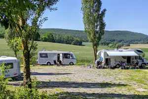 Wohnmobile auf einem Campingplatz zwischen Bäumen, mit Vorzelten und Fahrrädern vor grünen Wiesen und Hügeln.